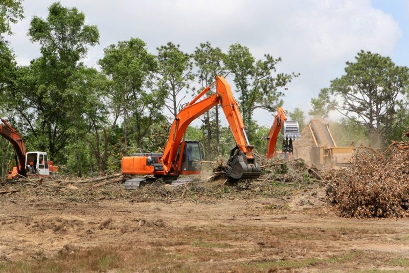 Orange excavators are busy with lot clearing, removing trees and brush in a forested area under a partly cloudy sky.